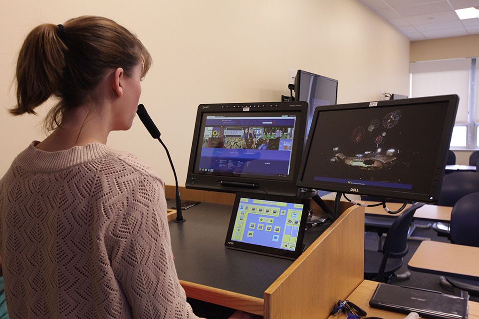 A female, back to the camera, standing at a classroom lectern set up with multiple monitors and a microphone.
